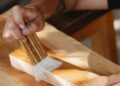 Manos aplicando barniz con brocha sobre una tabla de madera al barnizar madera en un taller